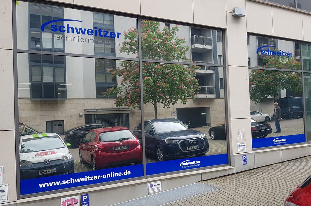 Schweitzer Geschäft auf Straße mit parkenden Autos, Fenstergrafiken und Bäumen im Hintergrund.