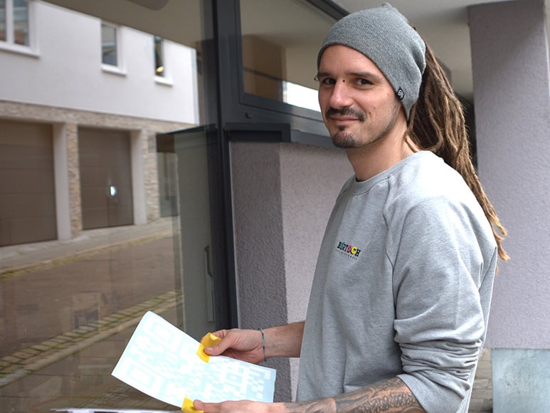 Mann mit Dreadlocks lächelt, trägt grauen Pullover und Mütze, hält gelbe Werkzeuge.