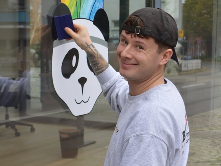 Ein lächelnder Mann mit Kappe malt einen bunten Panda auf eine Fensterscheibe.